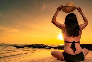 vue arrière d'une femme dodue asiatique portant un bikini et une main tenant un chapeau de paille assis sur la plage de sable se détendre et profiter des vacances à la plage paradisiaque tropicale. femme regarde le beau coucher de soleil. vacances d'été. ambiance estivale. photo