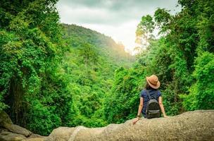 une femme heureuse voyage seule dans les bois. femme active de bonne humeur s'asseoir sur la pierre dans la vallée verte avec un arbre dense dans la forêt. bonnes vacances. vue arrière d'une femme heureuse portant un sac à dos, une bande intelligente et un chapeau. photo