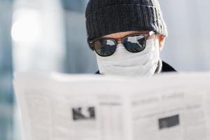 gros plan d'un homme adulte porte des lunettes de soleil, un chapeau et un masque médical stérile, lit le journal, découvre des nouvelles sur la situation dans le monde, la propagation du coronavirus, pose à l'extérieur sur un arrière-plan flou photo