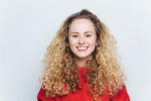 photo intérieure d'une femme blonde bouclée heureuse avec un sourire agréable et une expression joyeuse, vêtue d'un pull rouge chaud, modèles sur fond blanc. concept de personnes, de bonheur et d'émotions positives