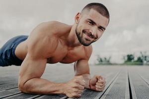 beau sportif barbu se tient en planche et fait du sport en plein air, s'entraîne régulièrement et travaille sur les muscles, démontre ses biceps, essaie de faire de son mieux. l'homme fait de l'exercice. photo