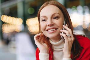 photo de tête d'une jeune femme séduisante avec une expression faciale heureuse, parle par téléphone portable, a les cheveux bruns, heureuse d'entendre des nouvelles, a du temps libre, arrière-plan flou avec espace de copie pour votre promotion