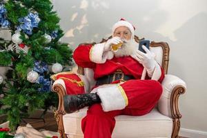 le père noël boit un verre de bière tout en utilisant son téléphone portable. temps de repos. boisson alcoolisée pendant les vacances. boire avec modération. bière artisanale. joyeux Noël. photo