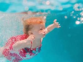 petite fille souriante en jolie robe moderne plongeant sous l'eau dans la piscine bleue. mode de vie actif, cours de natation pour enfants avec les parents. activité de sports nautiques pendant les vacances d'été en famille dans la station photo