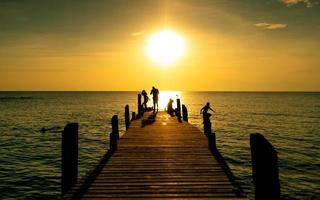 une famille heureuse joue ensemble pendant les vacances d'été au pont en bois au coucher du soleil. parent et enfants jouant à la plage en vacances. mer tropicale paradisiaque. les enfants s'amusent à sauter à la mer le soir. photo