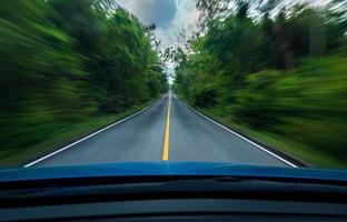 vue de face d'une voiture bleue conduisant à vitesse rapide au milieu d'une route goudronnée avec une ligne blanche et jaune de symbole de circulation dans la forêt verte. les arbres au bord de la route sont flous. voiture avec un mouvement de flou. photo