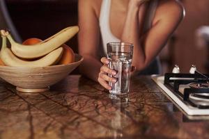 vue rapprochée de la fille tient le verre avec de l'eau. jeune femme debout dans la cuisine moderne près de la cuisinière à gaz photo