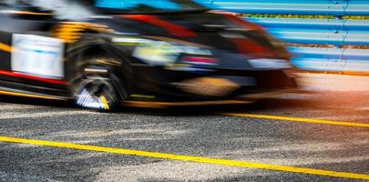 course de voitures de sport automobile sur route goudronnée avec clôture bleue et panneau de signalisation de ligne jaune. voiture avec conduite à vitesse rapide et mouvement flou. voiture de course noire à rayures rouges et jaunes. voiture sur circuit. photo