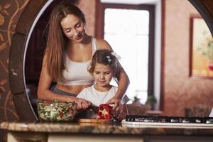 enfant apprend vite. jolie jeune femme debout dans la cuisine moderne près de la cuisinière à gaz et apprend à sa fille à préparer la nourriture photo