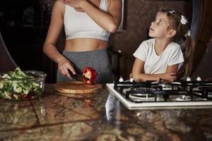 écoute la mère. jolie jeune femme debout dans la cuisine moderne près de la cuisinière à gaz et enseigne à sa fille comment préparer la nourriture photo