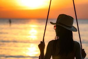 silhouette femme porter bikini et chapeau de paille balancer les balançoires à la plage pendant les vacances d'été au coucher du soleil. fille en maillot de bain s'asseoir sur des balançoires et regarder le beau coucher de soleil. ambiance estivale. femme voyage seule. photo