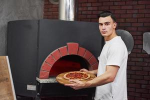 un boulanger élégant en vêtements blancs sort la pizza du four photo