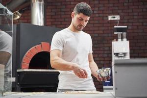 donner du solt. boulanger en chemise blanche jette des épices pour faire de délicieuses pizzas pour une commande au restaurant photo