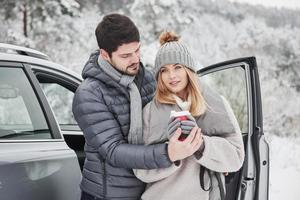 réchauffé par le café et les câlins en se tenant près de la voiture. un beau couple passe un week-end à la campagne en hiver photo