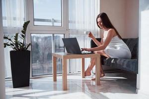 jolie jeune femme assise sur le canapé le matin dans sa chambre avec ordinateur portable photo