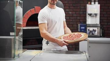 tenant dans des mains fortes. boulanger en chemise blanche avec pizza prête à être mise au four pour cuisiner photo