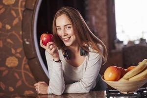 souriant en tenant la pomme dans la main. jolie jeune femme debout dans la cuisine moderne près de la cuisinière à gaz