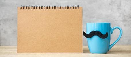 tasse à café bleue ou tasse à thé avec décor de moustache noire et bloc-notes en papier vide ou calendrier sur table. espace de copie vierge pour le texte. novembre bleu, bonne fête des pères et concept de journée internationale des hommes photo