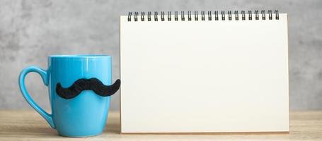 tasse à café bleue ou tasse à thé avec décor de moustache noire et bloc-notes en papier vide ou calendrier sur table. espace de copie vierge pour le texte. novembre bleu, bonne fête des pères et concept de journée internationale des hommes photo