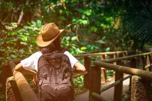 femme asiatique touriste avec chapeau et sac à dos debout et commencer à marcher sur le pont du sentier de la nature dans la forêt tropicale. seule jeune femme voyageuse en vacances en été photo