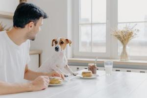 photo horizontale d'un homme et d'un chien mangeant ensemble, posent à la table de la cuisine contre une grande fenêtre, se regardent, ont de bonnes relations, profitent de l'atmosphère domestique. maison, animaux, concept de nutrition