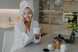 jolie femme souriante applique des patchs d'hydrogel sous les yeux aime se toiletter après la douche vêtue d'un peignoir boit du thé a une conversation téléphonique est assise sur une chaise à la maison. procédures de soins du visage photo