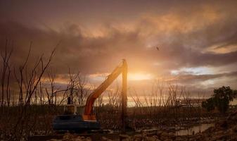 excavatrice draguant la boue à la mangrove. rétrocaveuse creusant la boue sur le chantier de construction. machine d'excavation enlevant les sédiments de la voie navigable. véhicule d'excavation. rétrocaveuse travaillant en soirée avec ciel coucher de soleil. photo