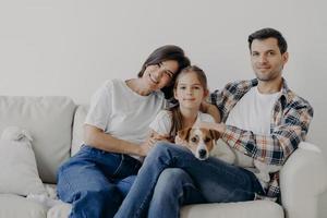 famille amicale pose ensemble au canapé, aime l'atmosphère domestique. le père, la mère, leur petite fille et leur chien de race passent le week-end à la maison, posent dans le salon, ont des expressions de visage heureux photo