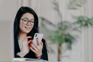 une femme d'affaires brune heureuse utilise un smartphone, porte des lunettes, envoie des SMS à son partenaire, travaille sur un projet, pose contre un intérieur confortable sur le lieu de travail. technologie, profession et concept de travail