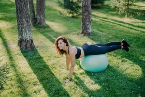 photo en plein air d'une femme brune active exerce avec un ballon de fitness pose sur l'herbe verte, vêtue de vêtements de sport, profite d'une journée ensoleillée et de l'air frais dans le parc, reste en forme, fait des exercices de gymnastique.