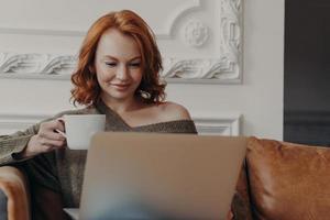 belle femme européenne rousse concentrée sur un ordinateur portable, réfléchit à l'accomplissement des tâches de travail, boit du café aromatique, s'assoit sur un canapé confortable, vérifie la base de données, analyse les informations photo