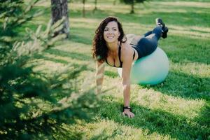 une femme brune souriante et heureuse a des exercices de yoga avec des poses de ballon de fitness au parc avec de l'herbe verte, a une expression joyeuse, reste en forme et en bonne santé, mène une vie active, a de la gymnastique le matin
