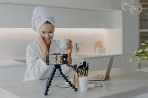 femme blogueuse beauté tire sur le vlog cosmétique regarde la caméra du smartphone parle avec les adeptes applique des patchs de collagène sous les yeux boit du thé porte un peignoir doux blanc enregistre la diffusion vidéo. photo