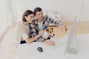 une femme et un homme ravis ont l'air heureux, regardent ensemble un film en ligne à la maison, jouent avec des points, s'assoient à une table blanche, recherchent des informations via le wifi, vêtus de vêtements décontractés, un couple familial navigue sur Internet photo