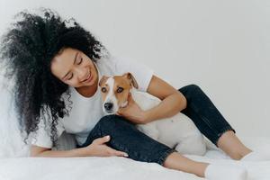 photo horizontale d'une jolie femme bouclée embrasse son chien préféré, porte un t-shirt blanc, un jean et des chaussettes, profite du temps ensemble, s'assoit sur le lit. bon début de journée. dame gaie joue avec un animal de compagnie dans la chambre