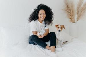 une femme afro-américaine séduisante et détendue boit une boisson chaude aromatique dans une tasse blanche, pose sur le lit avec un chien jack russell terrier, profite de l'atmosphère domestique, étant dans une chambre confortable à la maison photo