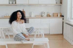 photo horizontale d'une femme à la peau sombre ravie qui rit agréablement, boit du café, regarde par la fenêtre dans la cuisine, vêtue de vêtements à la mode. femme souriante avec une boisson chaude savoureuse, se détend à la maison