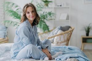 photo intérieure d'une jolie femme aux cheveux noirs assise les jambes croisées sur le lit, vêtue de vêtements de nuit, de chaussettes blanches, regarde directement la caméra, se sent détendue tout en écoutant de la musique calme dans les écouteurs. repos et congé