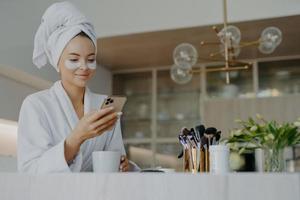 photo d'une jeune femme séduisante porte une serviette de bain sur la tête et une robe de chambre blanche douce tient un téléphone portable aime la communication en ligne applique des patchs hydratants sous les yeux boit une boisson chaude