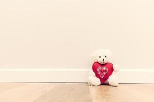 adorable ours en peluche avec un gros coeur en peluche rouge. assis sur un plancher en bois contre un mur blanc. photo