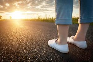 femme en baskets blanches debout sur la route goudronnée vers le soleil. voyages, concepts de liberté. photo