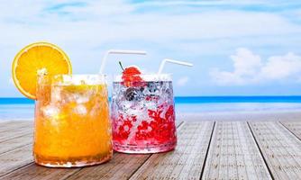 jus d'orange dans un verre transparent avec des glaçons et du soda à la fraise mocktail ne pas mélanger l'alcool. fraises fraiches. boissons fraîches posées sur le balcon en bois au bord de la mer en milieu de journée. rendu 3d photo
