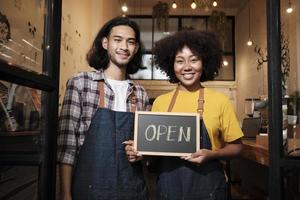 deux jeunes partenaires de barista en démarrage avec des tabliers se tiennent à la porte d'un café décontracté, des lettres à bord et montrent un signe ouvert, des sourires heureux et joyeux avec des emplois de service de café et de nouveaux entrepreneurs. photo