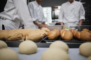 gros plan et mise au point sélective sur des baguettes et du pain délicieux devant l'équipe de chefs en uniforme blanc, pétrir la pâte à pâtisserie crue, préparer des aliments de boulangerie frais, cuire au four dans la cuisine du restaurant. photo