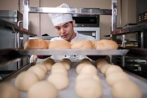 jeune chef masculin asiatique professionnel en uniforme de cuisinier blanc avec chapeau, gants et tablier faisant du pain à partir de pâte à pâtisserie, préparant des aliments de boulangerie frais, cuisant au four dans la cuisine en acier inoxydable du restaurant. photo