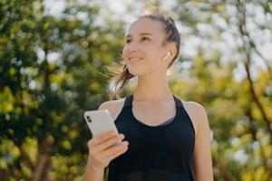 une femme joyeuse et réfléchie avec une queue de cheval a marché en plein air écoute de la musique tout en faisant du sport détient un smartphone moderne mène un mode de vie sain axé sur des poses à distance au parc contre des arbres photo