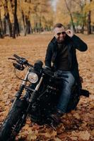 image d'un motocycliste masculin joyeux sourit joyeusement, garde la main sur les nuances, s'assoit sur un vélo rapide, s'amuse en plein air, aime voyager en moto, passe du temps libre dans le parc d'automne. notion de voyage. photo