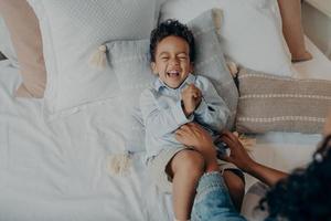 adorable petit fils allongé sur des oreillers dans son lit et riant pendant que maman le chatouillait photo