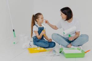 une mère et une fille positives et amicales se badigeonnent de peinture blanche, choisissent la couleur de la palette, tiennent des rouleaux de peinture, s'assoient par terre dans leur appartement aux murs blancs. salle de peinture dans une maison neuve photo