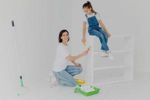 photo horizontale d'une mère attentionnée et de sa fille occupées à rénover des meubles à la maison, à utiliser de la peinture blanche et un pinceau, un rouleau, un travail de finition presque heureux, à peindre une nouvelle maison, habillés avec désinvolture